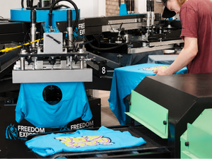 Person working with a teal t-shirt on a printing machine in a workshop setting.